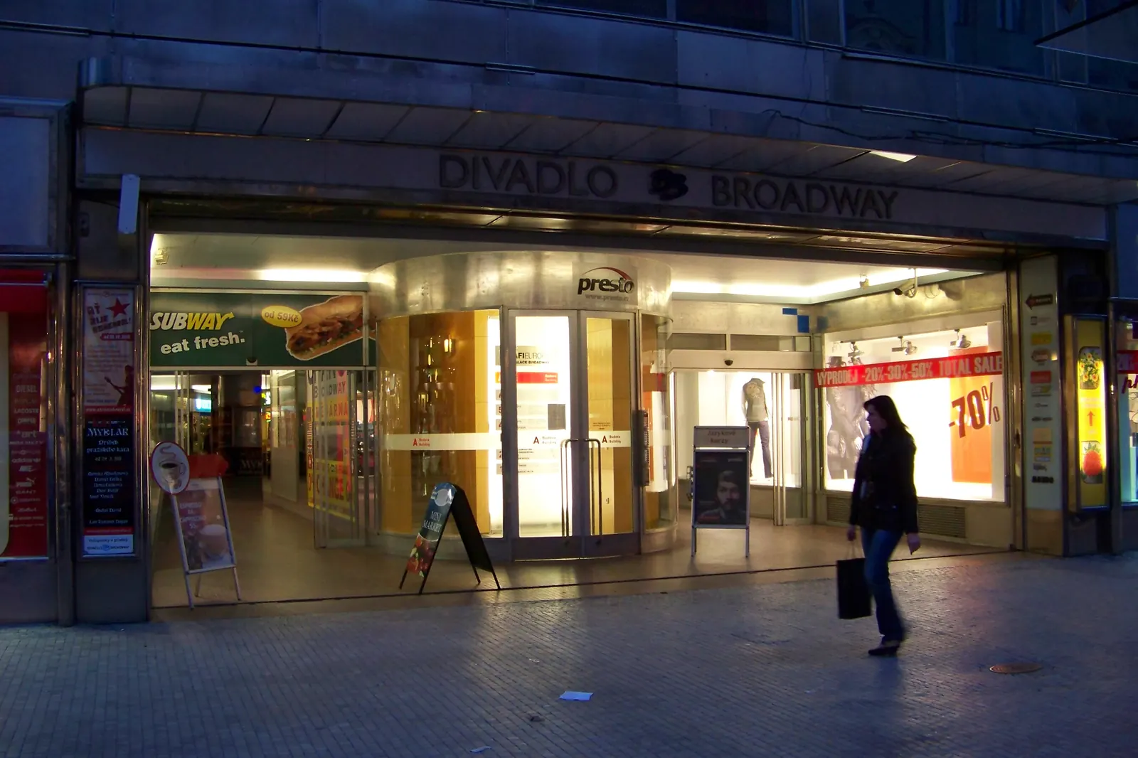 The entrance to the Broadway arcade on Na Příkopě in Prague at dusk, a glass-fronted passage with lit shop windows under a sign reading Divadlo Broadway