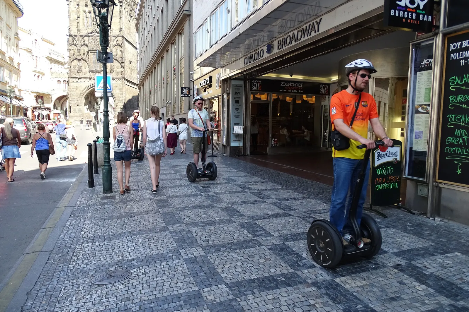 The Celetná end of the Broadway arcade, with the Powder Tower rising behind a cobbled street where people walk and a man rides a two-wheeled scooter