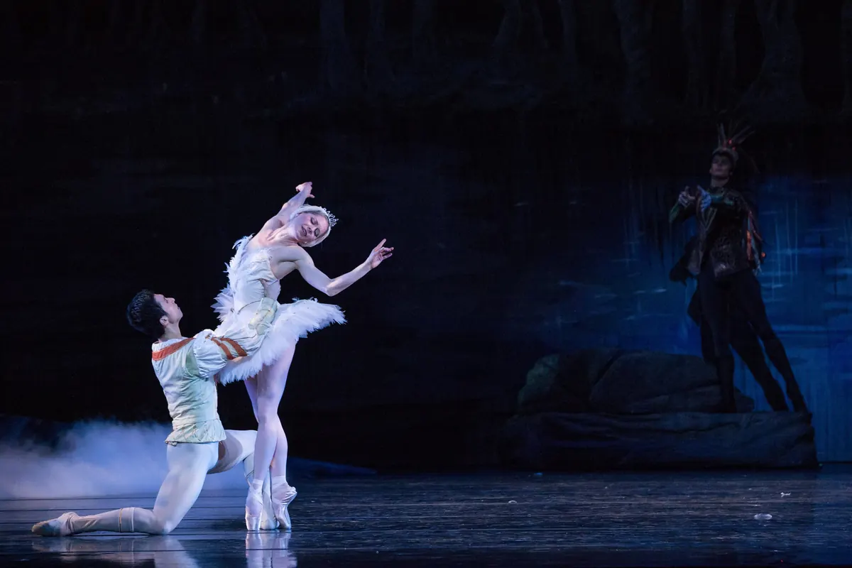 A male dancer in a pale doublet kneels on a misty blue stage and lifts a ballerina in a white feathered tutu and swan headpiece, with a dark figure in the background