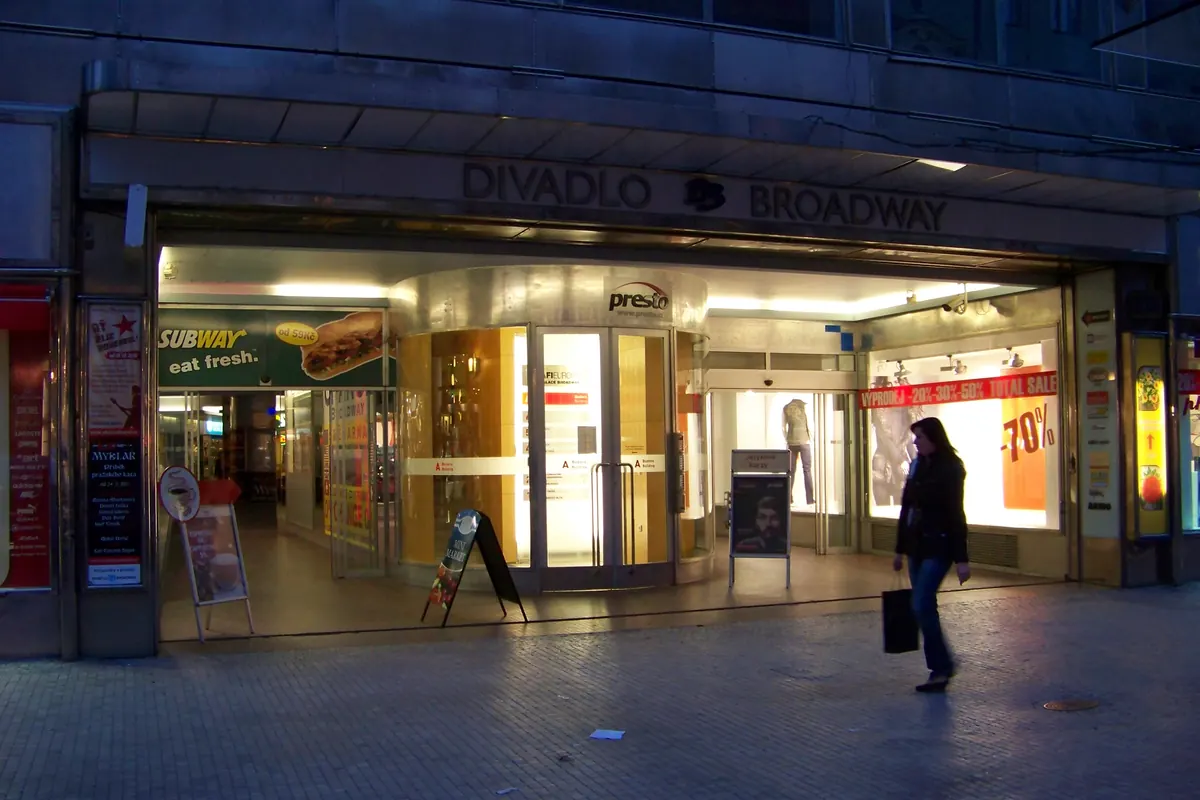 The glass-fronted entrance of the Broadway arcade on Na Příkopě at dusk, with lit shop windows and a sign above reading Divadlo Broadway