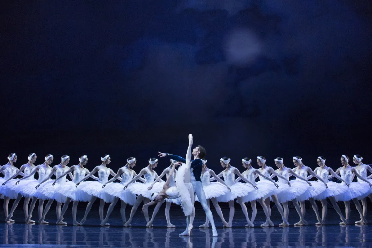 A long line of dancers in white tutus stands in a row across a dark blue stage, with one ballerina in front of them in a lifted pose