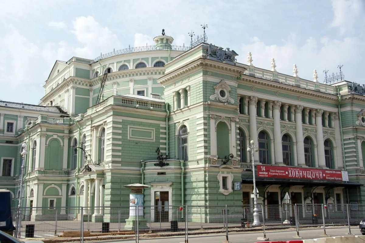 A pale green neoclassical theatre building with white columns, a domed roof and a red banner above the entrance, seen from the street