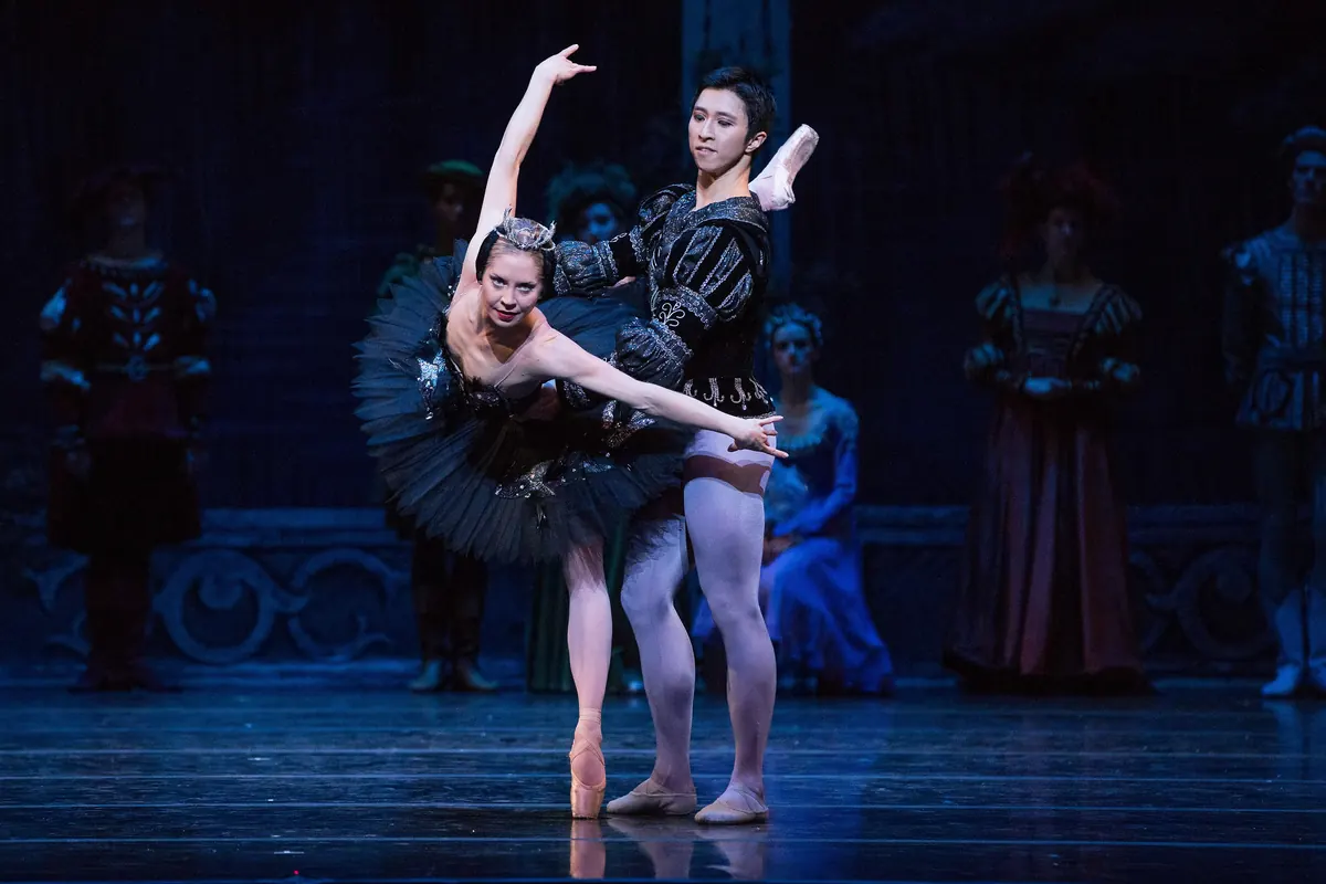A ballerina in a black tutu leans into a deep pose beside a male dancer in a dark doublet on a blue-lit stage, with courtiers in period dress standing behind