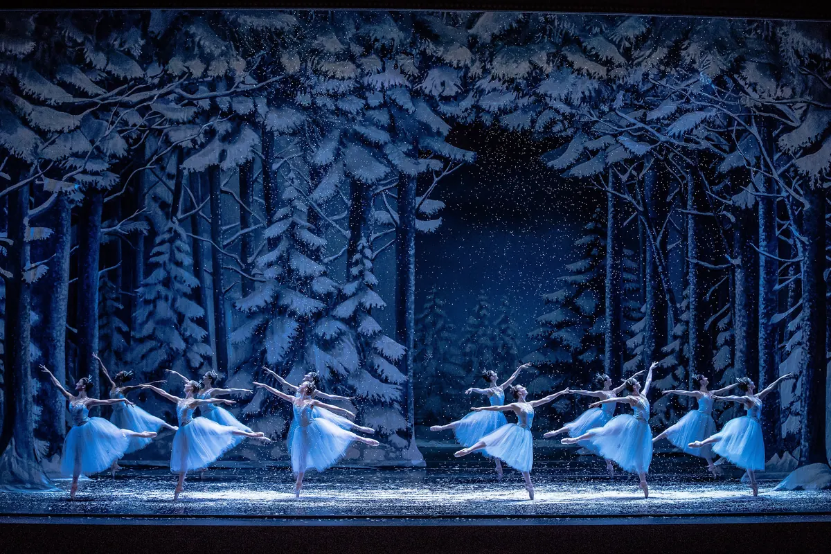 Dancers in pale blue-white tutus stand in lines on a dark stage in front of a painted forest of snow-covered fir trees, with snow falling